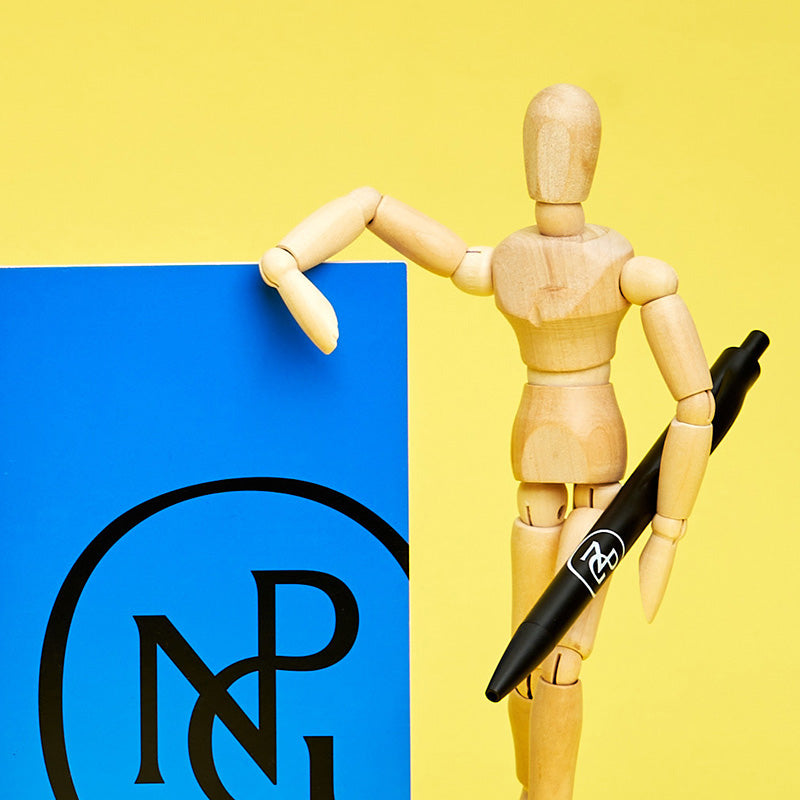 A small wooden mannequin holding a black npg pen and leaning on a blue npg book.