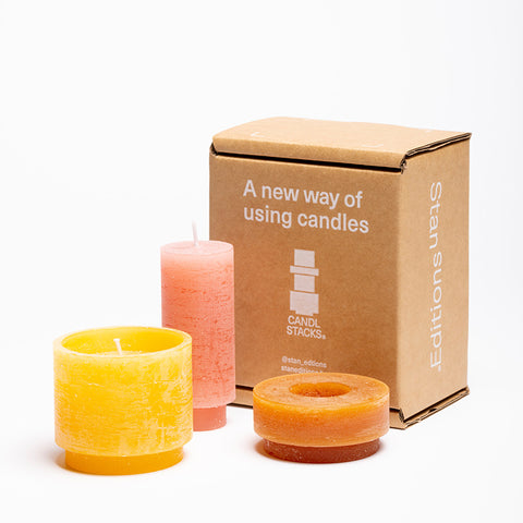 Three stackable candles, unstacked, in red, orange and yellow with its packaging. 
