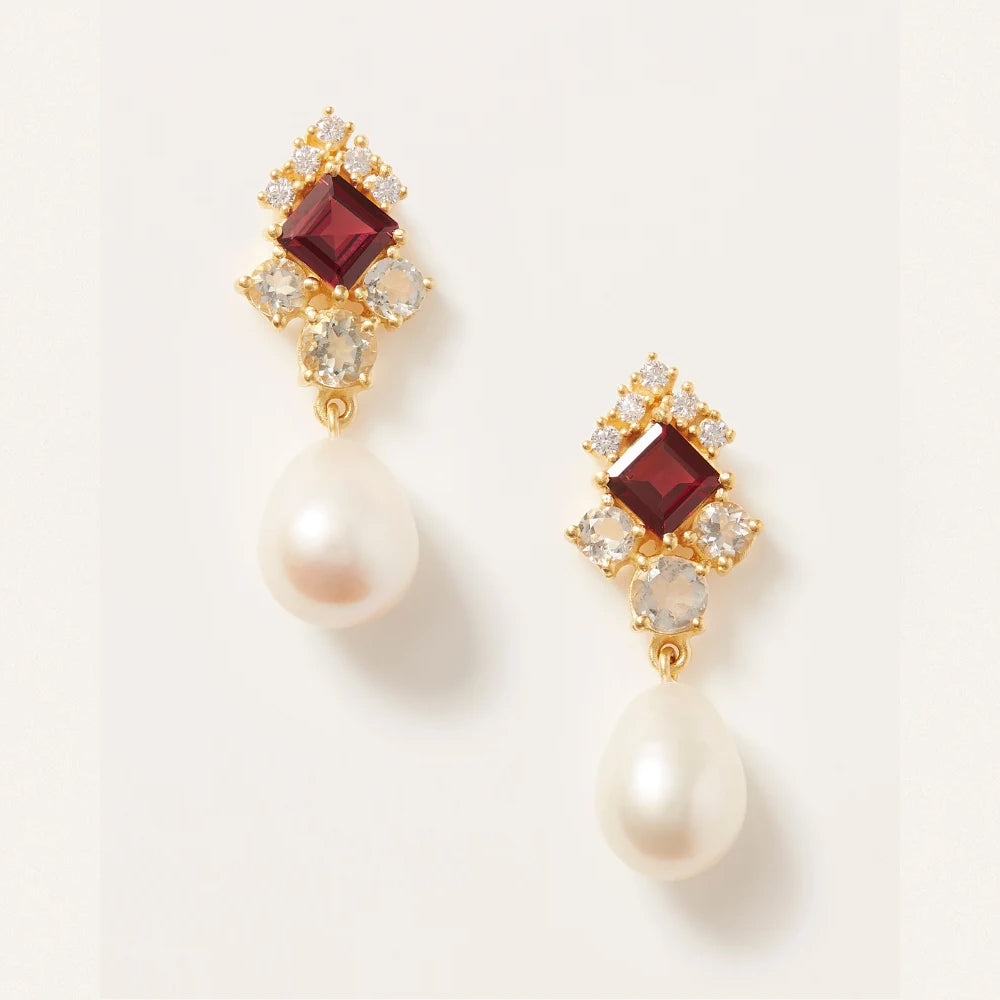 A pair of gold drop earrings featuring a square dark red gemstone, small clear crystals, and a teardrop shaped white pearl dangling below, set against a clean off white background.