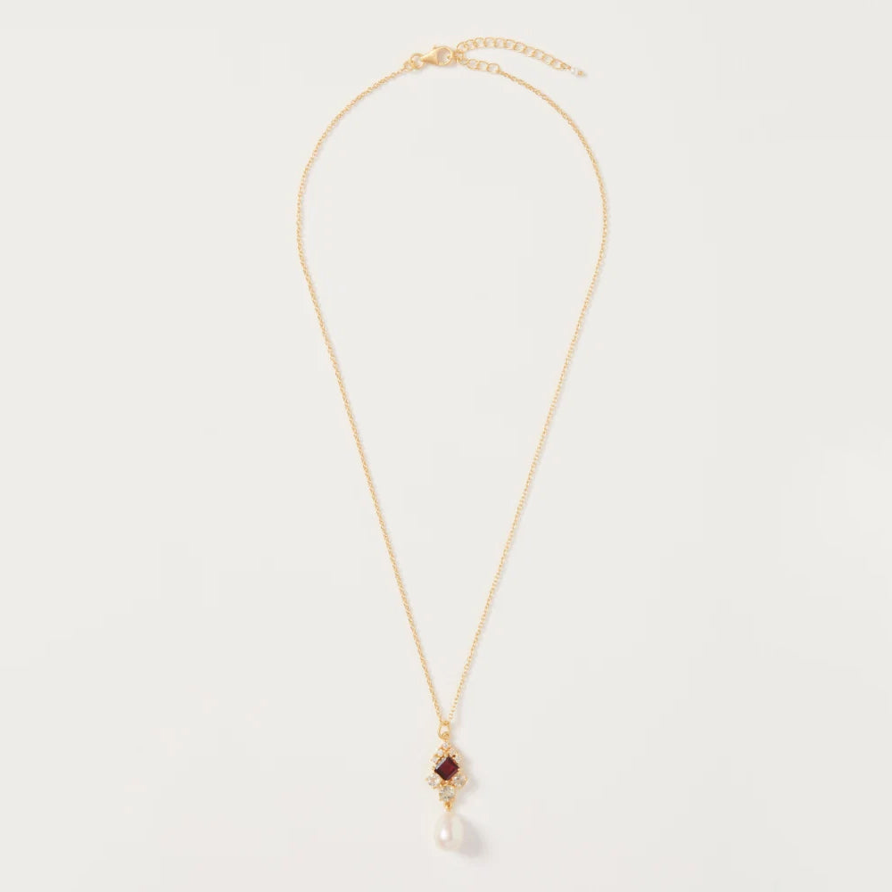 A delicate gold chain necklace featuring a small diamond shaped pendant with a dark red gemstone and a dangling white pearl, displayed on a clean off white background.