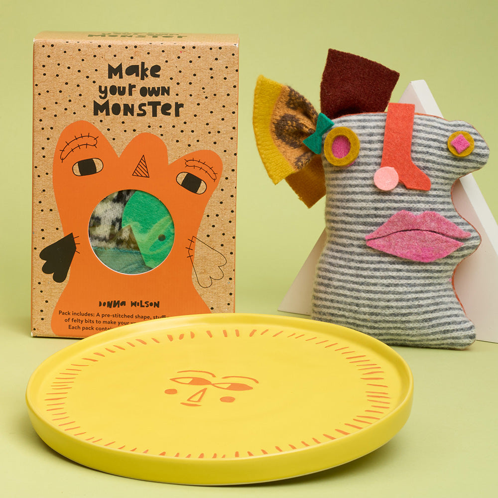 Make your own monster kit box next too a plush monster toy and a yellow ceramic sun design plate against green background.