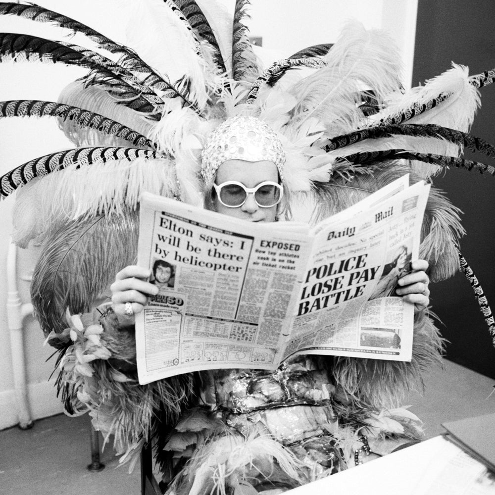 Close up of a man in feathered costume and white framed glasses, reading a newspaper.