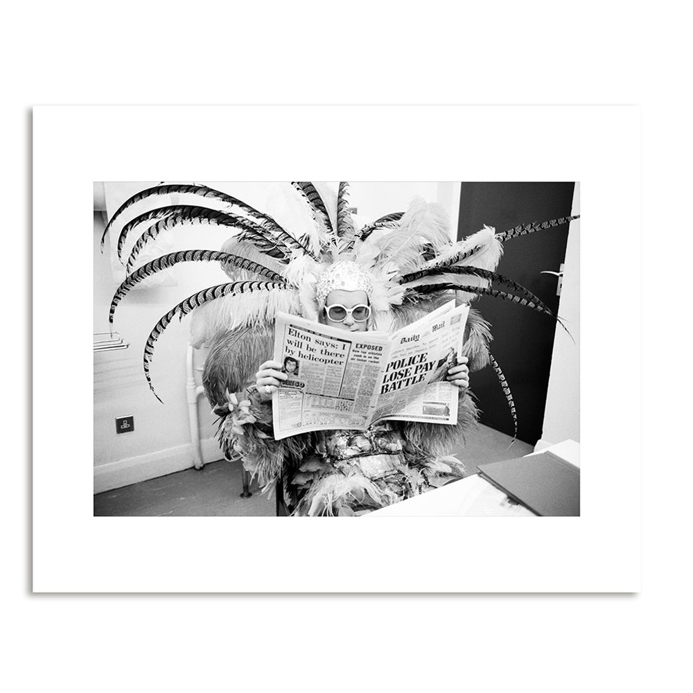 A black and white photograph of a man sitting in a flamboyant feathered costume reading a newspaper.