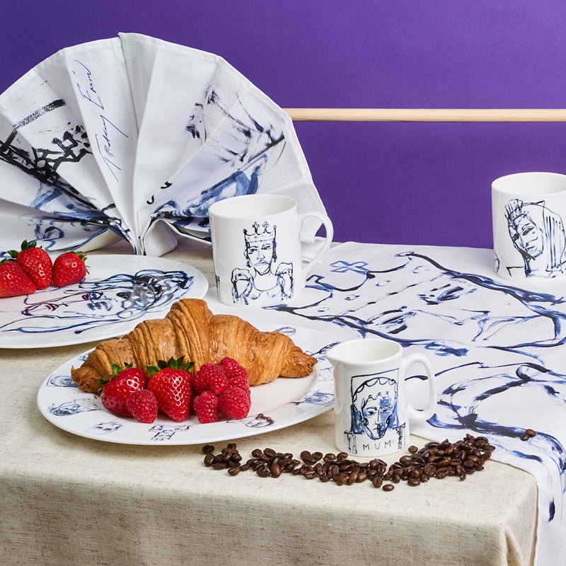 Table setting with croissants, berries, coffee beans, and mugs on a tablecloth with artistic designs.
