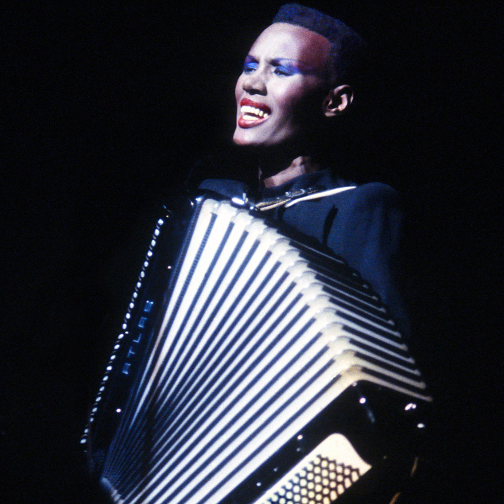 A close up photograph of woman in a black dress who sings and plays an accordion against a black background.
