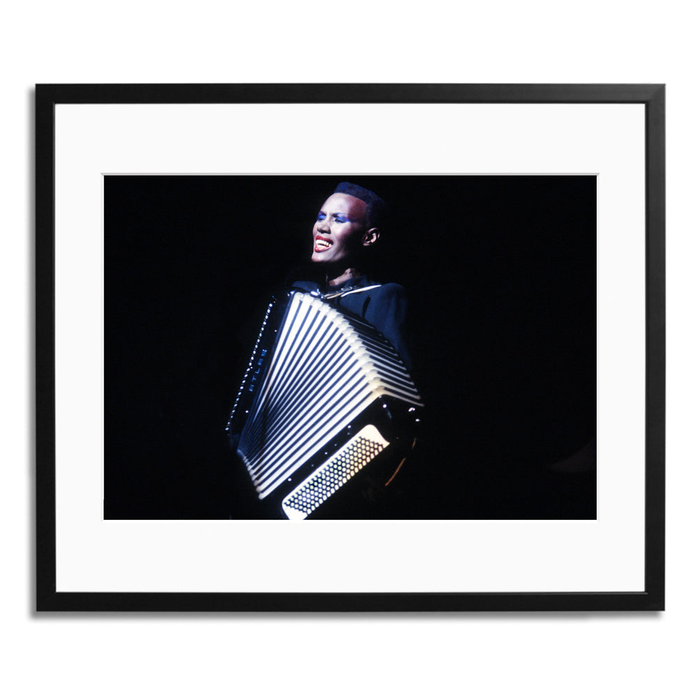 A close up photograph in a black frame of woman in a black dress who sings and plays an accordion against a black background.