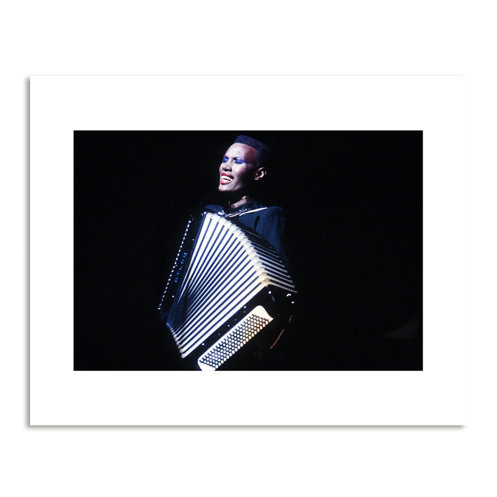 A woman in a black dress sings and plays an accordion against a black background.
