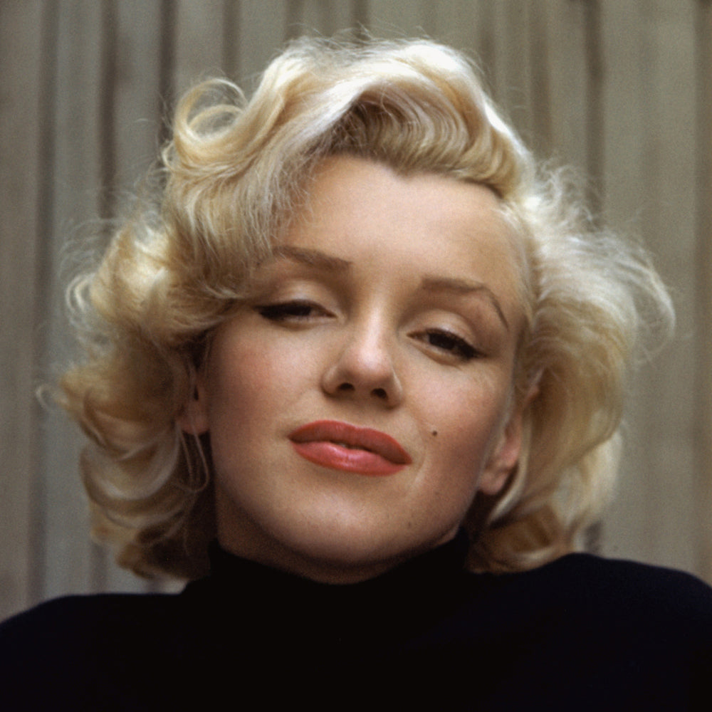 A close up photograph of a women with short blonde hair and a high neck black jumper with a fence in the background.