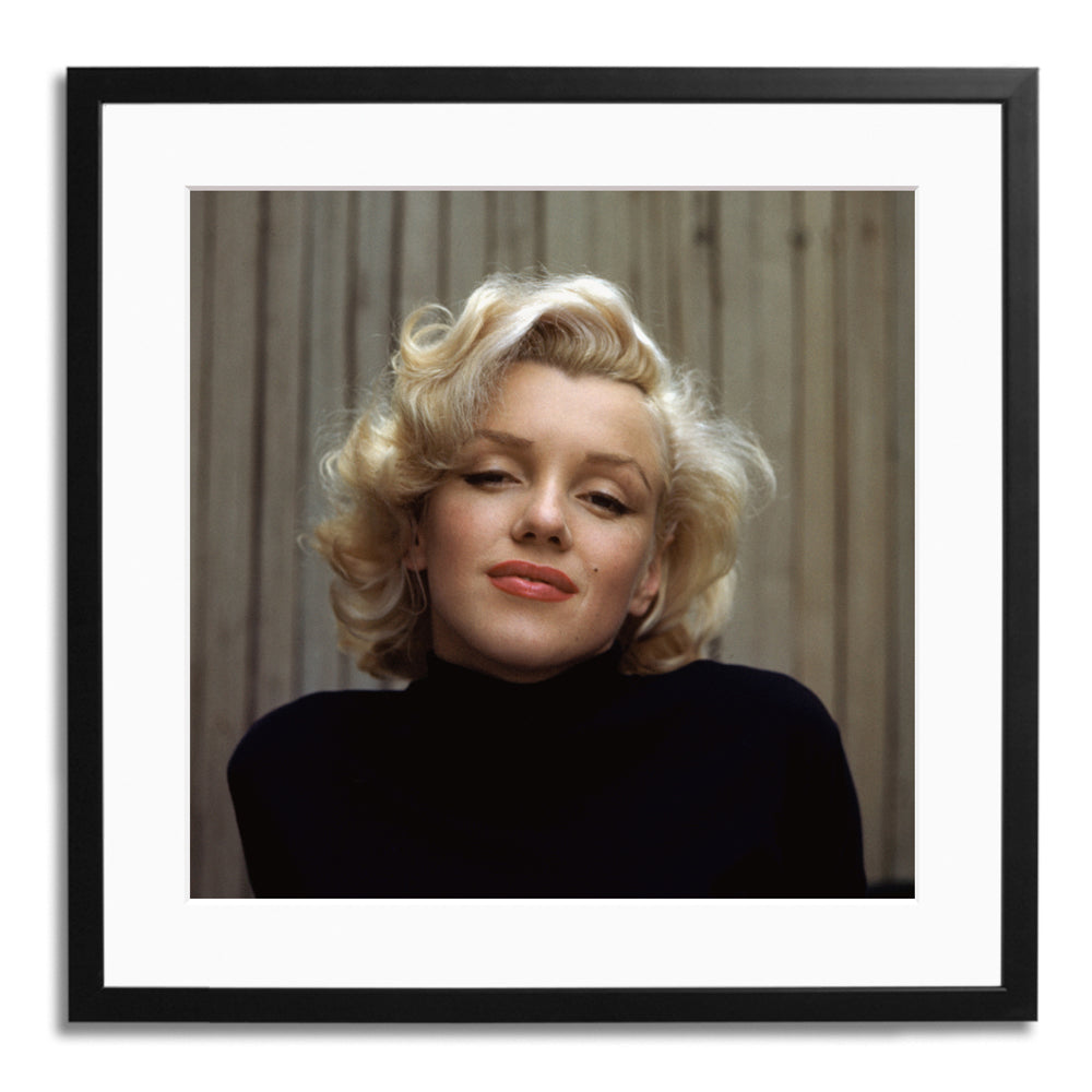 A close up photograph in a black frame of a women with short blonde hair and a high neck black jumper with a fence in the background.