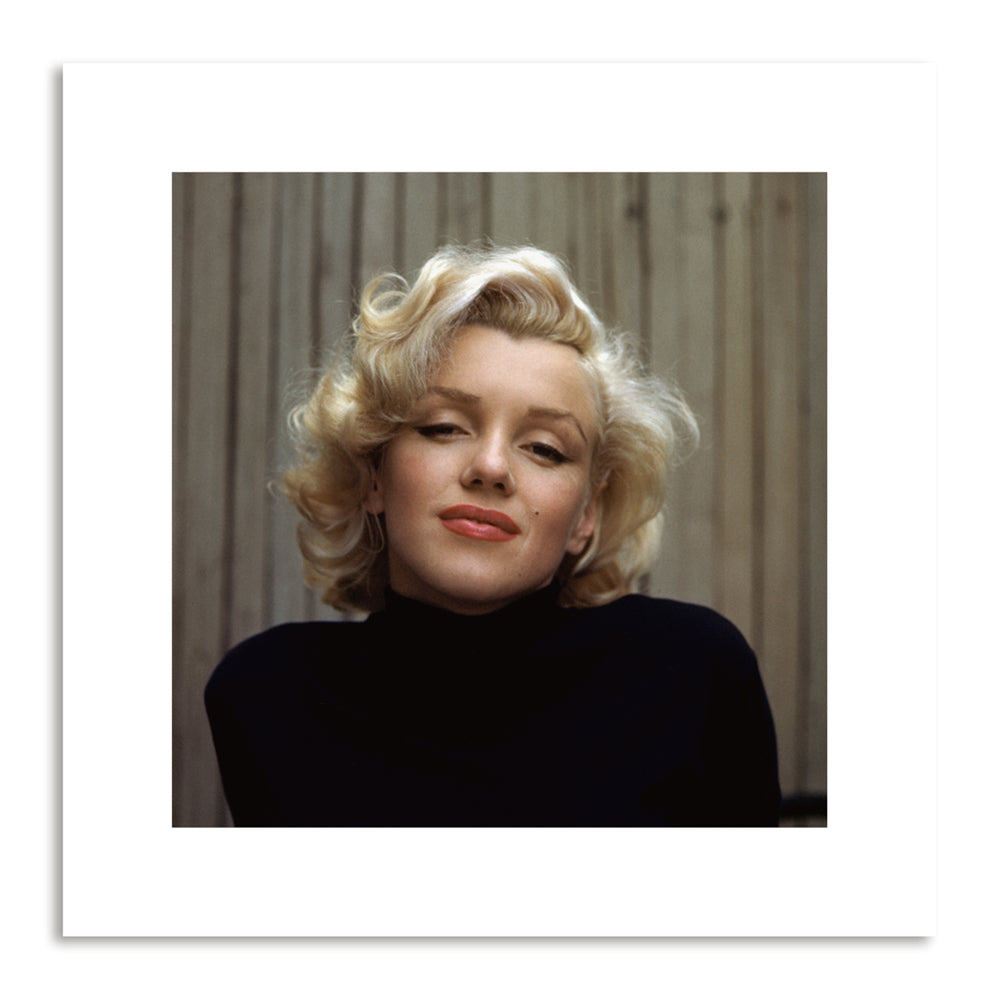 A close up photograph of a women with short blonde hair and a high neck black jumper sits in front of a wooden fence.