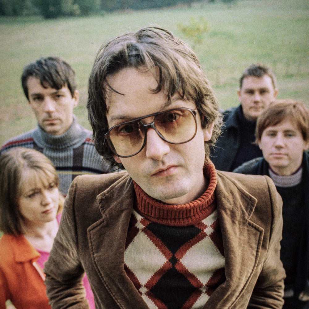 A close up of a photograph of 5 people in a field, in the foreground a man in a diamond print jumper and glasses poses with his hands in his pockets looking in to camera.
