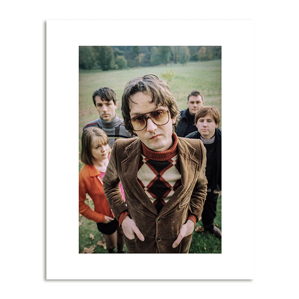 A photograph of 5 people in a field, in the foreground a man in a diamond print jumper and glasses poses with his hands in his pockets looking in to camera.