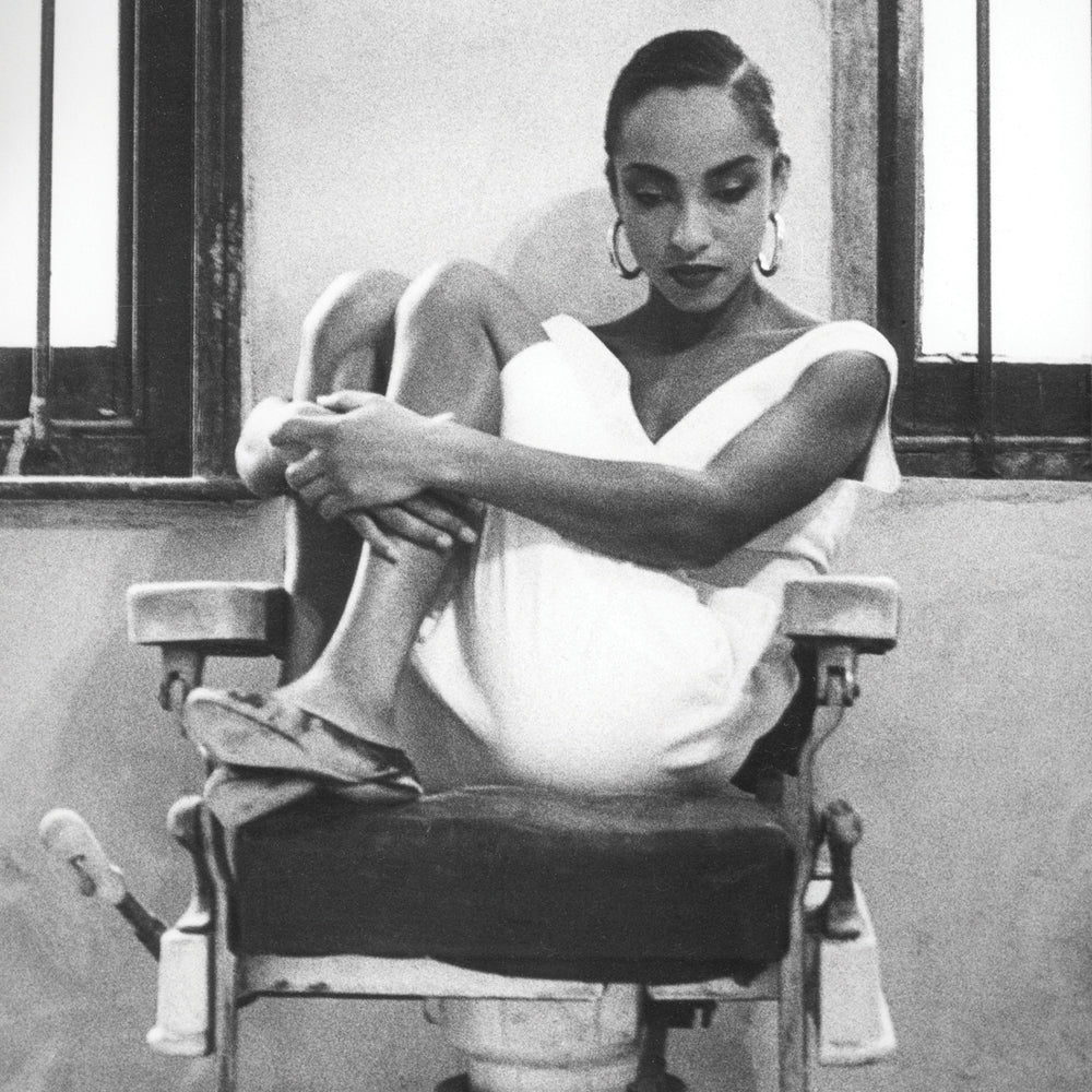 Close up of a black and white photograph a women sits on a chair in a white dress with her knees pulled in to her chest looking down to the floor.