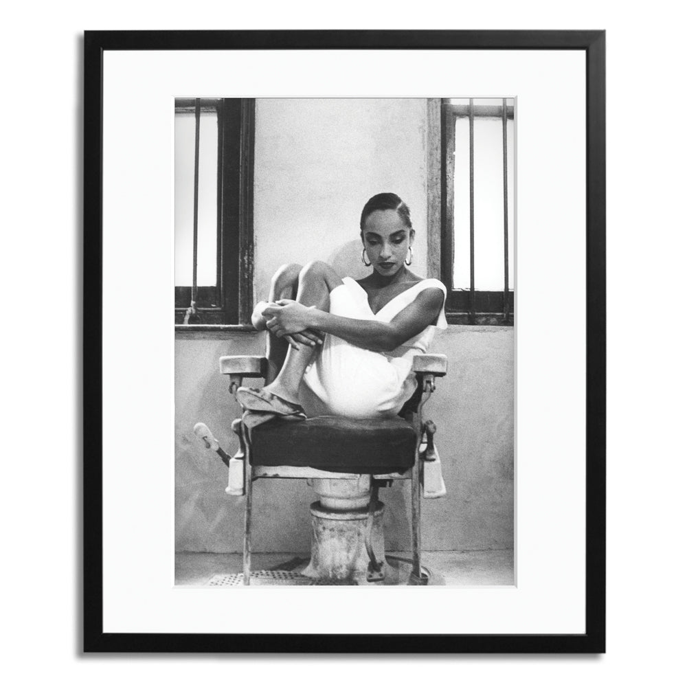 A black and white photograph in a black frame a women sits on a chair in a white dress with her knees pulled in to her chest looking down to the floor.
