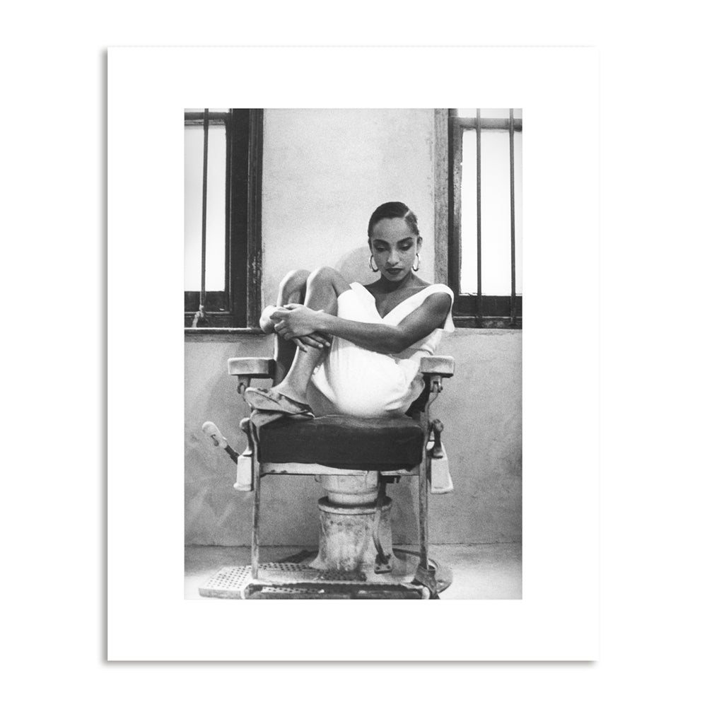 Black and white photograph a women sits on a chair in a white dress with her knees pulled in to her chest looking down to the floor.