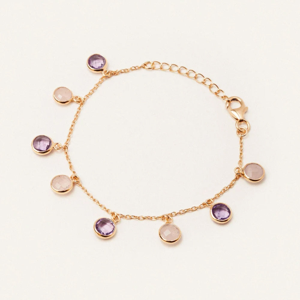 A delicate gold chain bracelet with seven round bezel set charms, including four purple amethyst stones and three pale pink stones, displayed on a clean off white background.