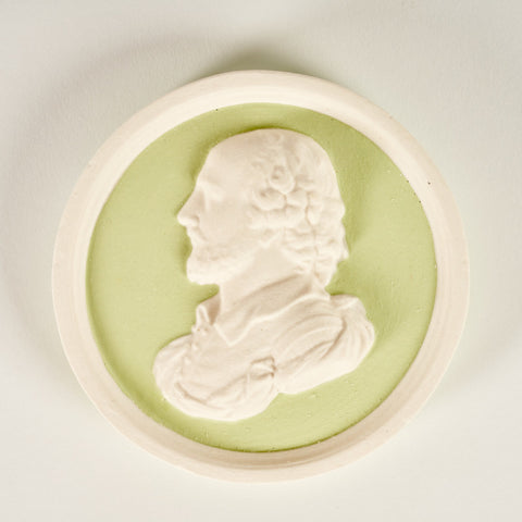A round plaque in green and white featuring a raised side profile portrait of William Shakespeare.