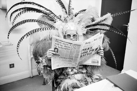Black and white photograph of Elton John in costume reading a newspaper, taken by Terry O'neill, 1977.