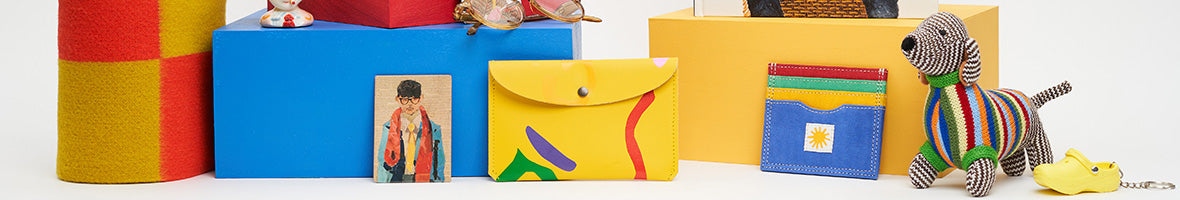 David hockney exhibition range featuring a yellow popper purse, self portrait magnet and knitted sausage dog against yellow and blue coloured blocks.