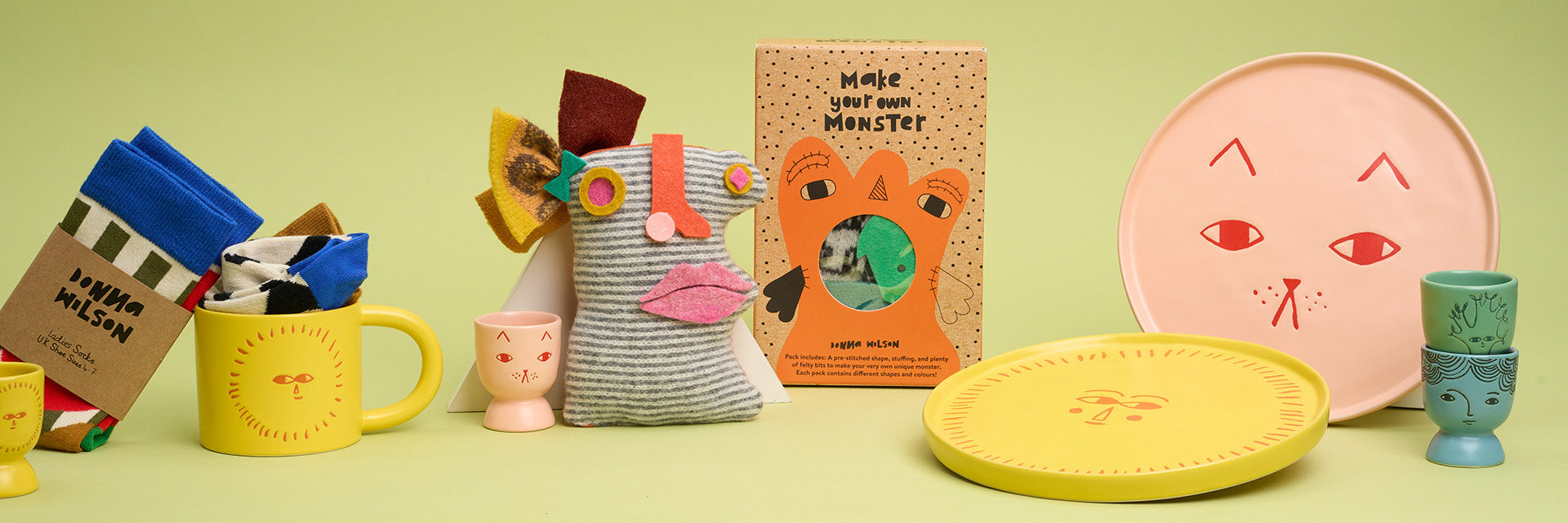 A display of pastel ceramic plates with illustrated faces, sock puppet and mug against a green background.