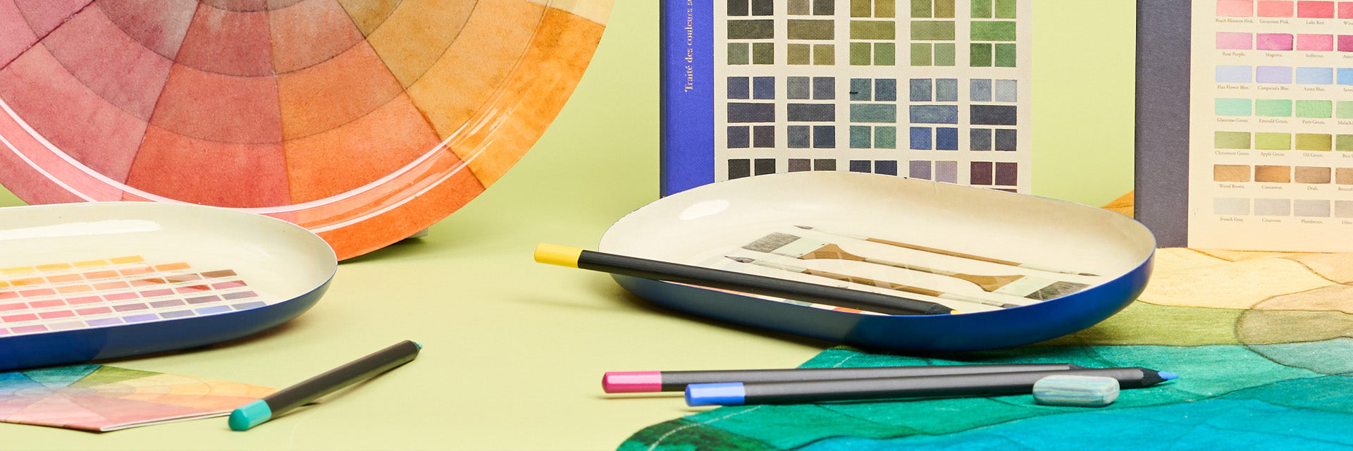 A display of colour swatch design sketchbooks and trays alongside pencils against a green background.