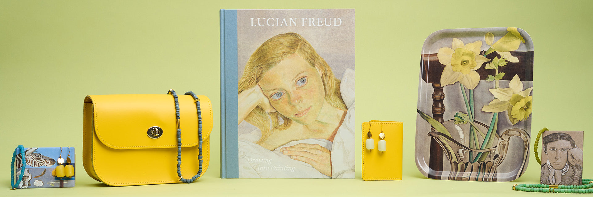 A lucian freud catalogue, daffodil tray, yellow satchel bag and jewellery are displayed alongside each other in a row against a green background.