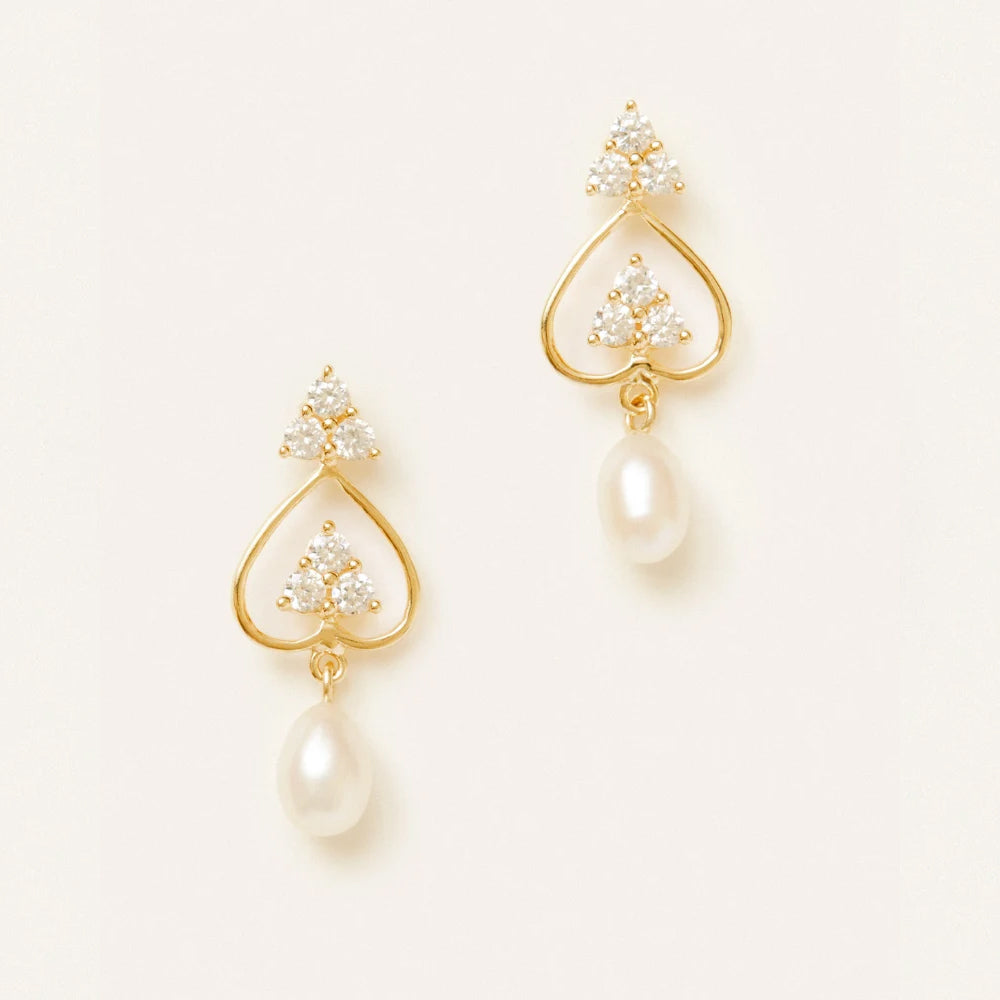 A pair of gold dangle earrings with clear crystal clusters at the top and a teardrop shaped white pearl hanging below, set against a plain off white background.