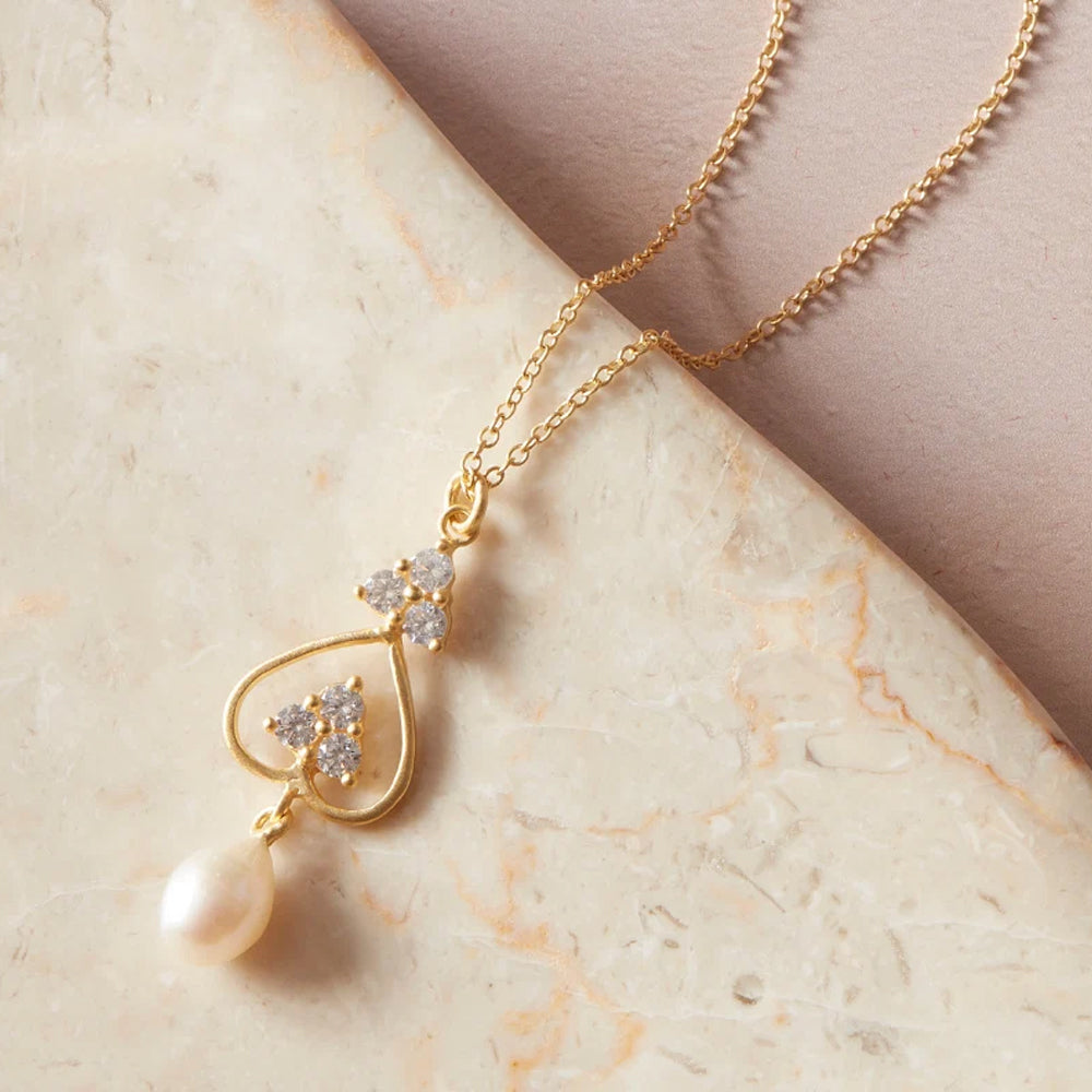 A gold pendant necklace with a heart shaped wire frame, small clear crystal clusters, and a dangling white pearl, displayed on a beige stone surface.
