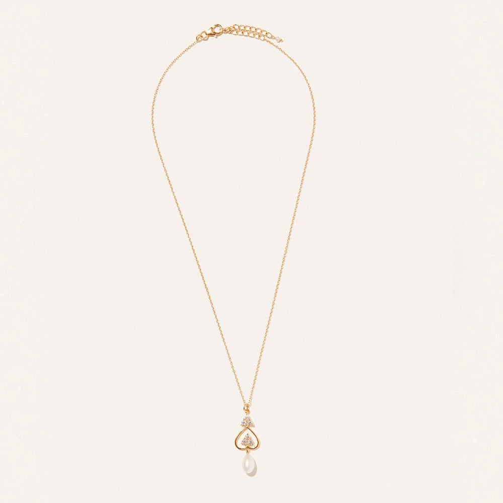 A delicate gold chain necklace with a small heart shaped pendant set with tiny clear stones and a dangling white pearl, displayed on a clean off white background.