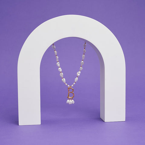 A pearl necklace with gold 'B' letter pendant is suspended from a white arch.