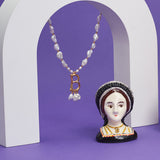 Small egg cup with a portrait of Anne Boleyn on it, in a black dress with her 'B' pearl necklace on. In the background, there is a 'B' pearl necklace hanging from a small arch.