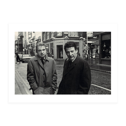 Black and white postcard of two men in overcoats in the middle of a busy road looking at the camera. 