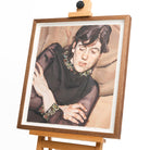 A framed print of a women in a black dress with her arms crossed, the framed rests upon a wooden easel.