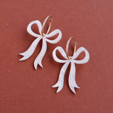 A pair of cream acrylic bow shaped earrings hanging from gold hoops. 