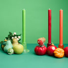 Three tall pillar candles in green, red, and orange displayed on a green surface with decorative autumn and shaped candles in the shape of pumpkins and pears