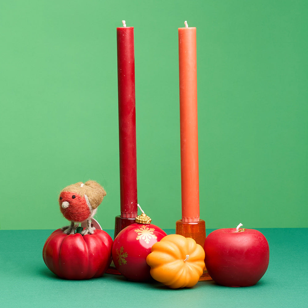 Two tall pillar candles in red and orange standing in a decorative arrangement with candles in the shape of a red apple, a small orange pumpkin, along with a felt bird on a green background.