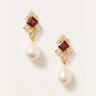 A pair of gold drop earrings featuring a square dark red gemstone, small clear crystals, and a teardrop-shaped white pearl dangling below, set against a clean off-white background.