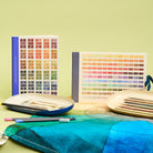 Colour swatches design sketchbook, journal and art supplies on a gradient colour tea towel against a green background.