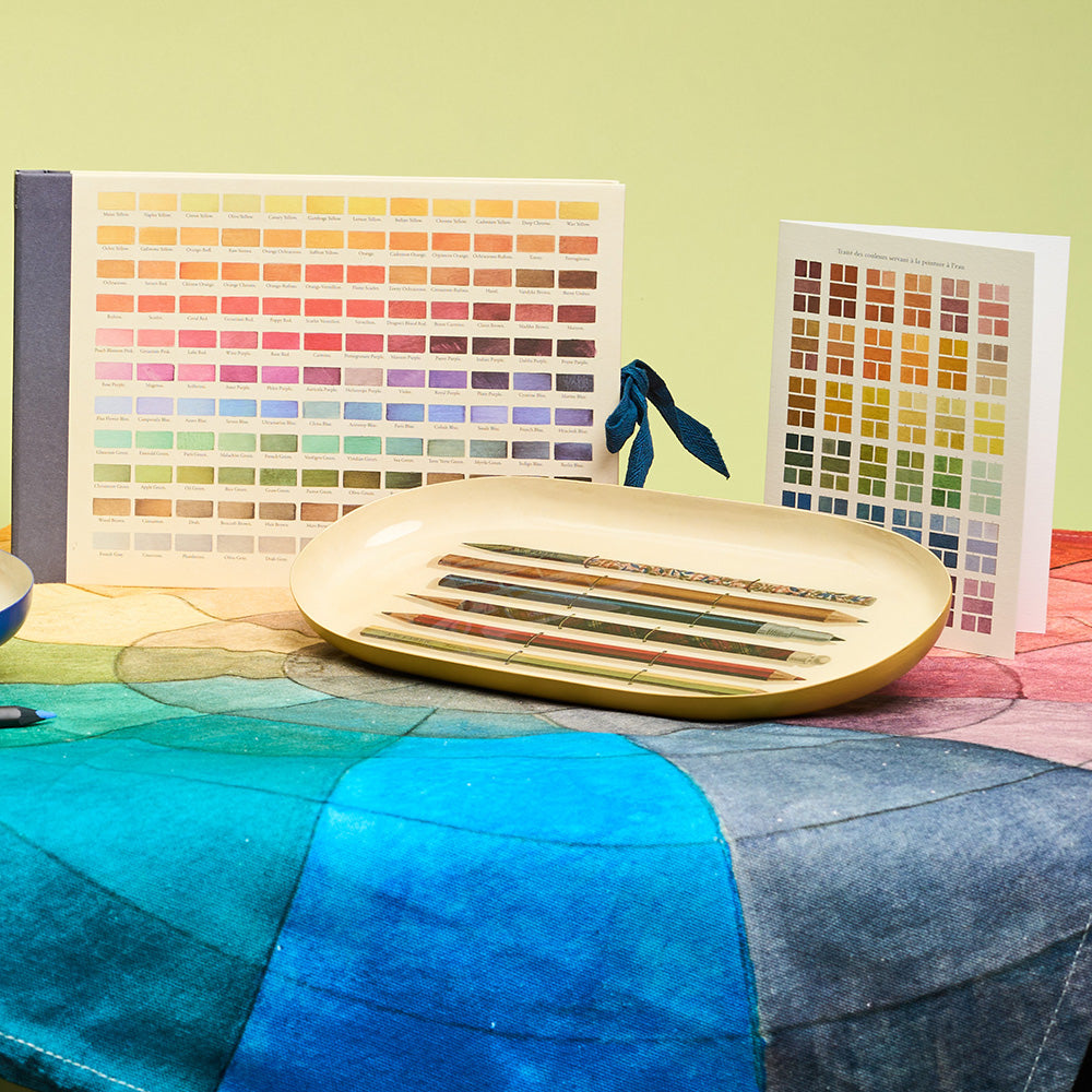 Colour swatch design sketchbook on a gradient colour tea towel and enamel tray with illustrated pencils against a green background.