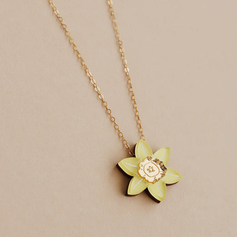 Acrylic yellow daffodil shaped pendant on a gold chain against a pale pink background.
