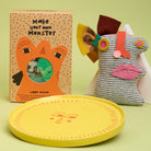Make Your Own Monster kit box next too a plush monster toy and a yellow ceramic sun design plate against green background.
