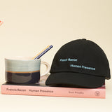 Black baseball hat on the pink exhibition catalogue with a blue-toned mug with some pencils sticking out. 