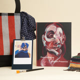 A collection of items, including a catalogue, a navy bandana with white polka dots on a tote bag with a clipboard and a postcard on it. 