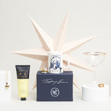 A series of small gift items including a jug, champagne saucer glass and handcream are displayed in front of a folded paper star decoration.