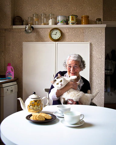 A portrait of a woman with white hair sat at a kitchen table holding her white cat.