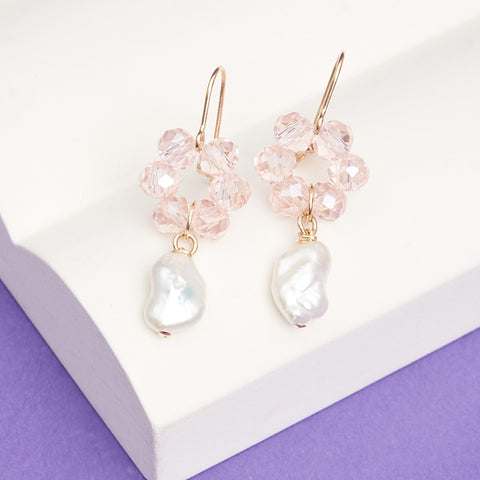 Dangling earrings with light pink glass beads in a flower shape with pearls hanging from the bottom.