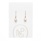 A pair of earrings featuring a single pearl hanging form a gold hook attached to NPG packaging.