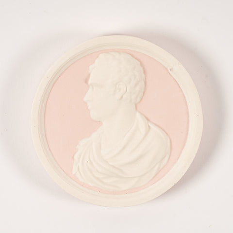 A round plaque in pink and white featuring a raised side profile portrait of Lord Byron.