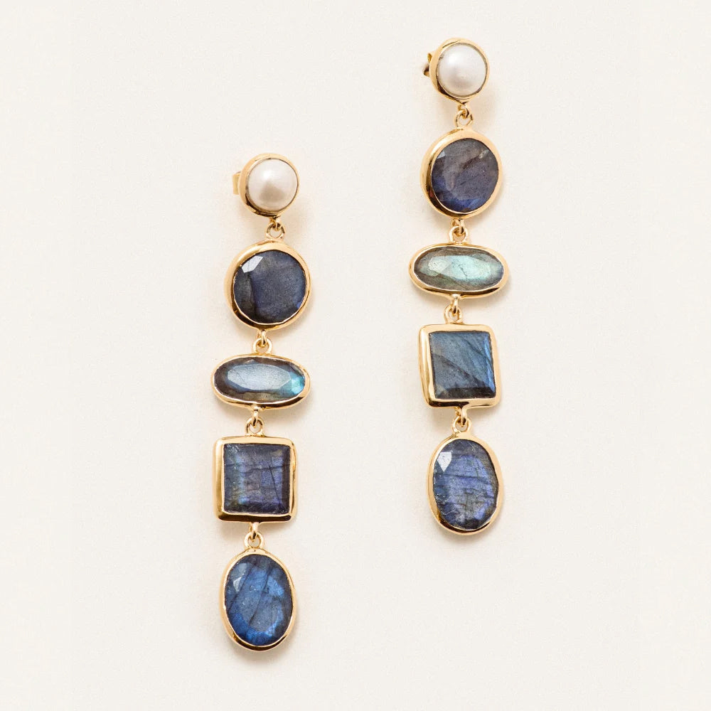 A pair of gold drop earrings featuring a white pearl at the top, followed by multiple blue labradorite stones in oval, round, and square settings, displayed on a clean off white background.