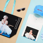 A selection of products including a tote bag and t-shirt with an image of a woman sitting in a tutu, hat and glasses against a blue and beige background.